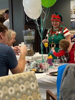 An elf is holding orange juice sitting next to a young toddler and family.