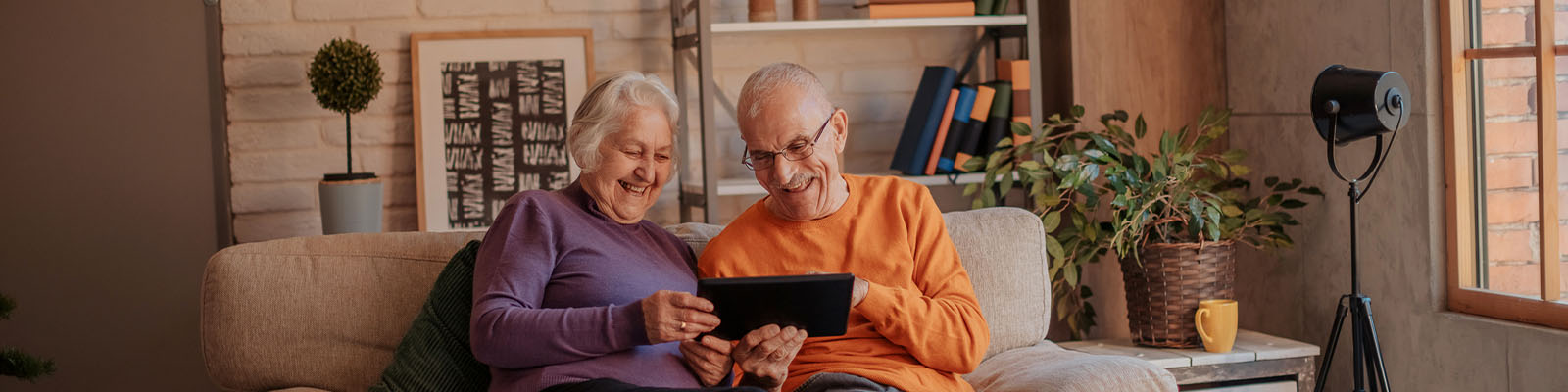 An elderly man and elderly woman sitting together on a grey sofa as they hold a black smart tablet and look down at it